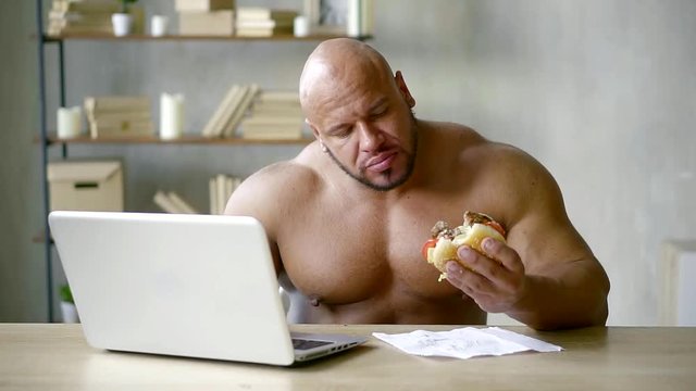 bald male bodybuilder with big muscles sitting at the table working on laptop and eating triple Burger with beef Patty
