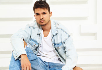 Portrait of handsome young model man dressed in jeans clothes sitting near white textured wall