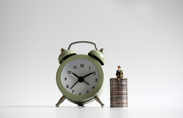 A miniature man sitting on a pile of coins by the alarm clock.
