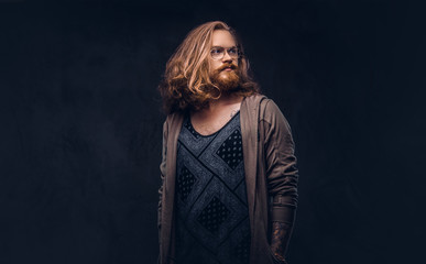 Close-up portrait of a redhead hipster male with long luxuriant hair and full beard dressed in casual clothes standing in a studio, looking away. Isolated on the dark background.
