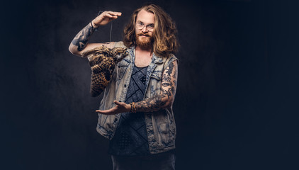 Portrait of a tattoed redhead hipster male with long luxuriant hair and full beard dressed in a t-shirt and jacket holds a keeps the scarecrow of an owl in a studio. Isolated on the dark background.