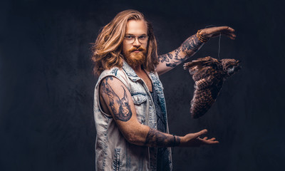 Portrait of a tattoed redhead hipster male with long luxuriant hair and full beard dressed in a t-shirt and jacket holds a keeps the scarecrow of an owl in a studio. Isolated on the dark background.