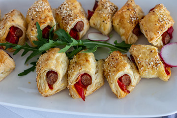 Snacks from puff pastry with sausage in the middle, sprinkled with sesame, decorated with arugula and radish close up 