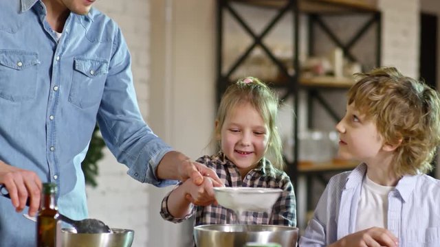 Medium shot of young father in denim shirt helping excited little girl sift flour into metal container when cooking with two children
