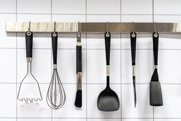 Set of black cooking utensils hanging on stainless bar on white modern ceramic wall tiles in the...