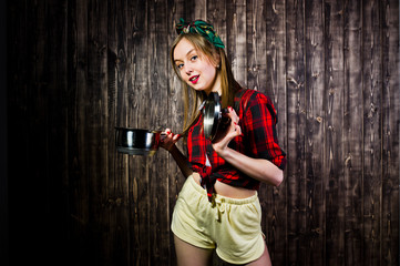 Fototapeta premium Young funny housewife in checkered shirt and yellow shorts pin up style with saucepan on wooden background.