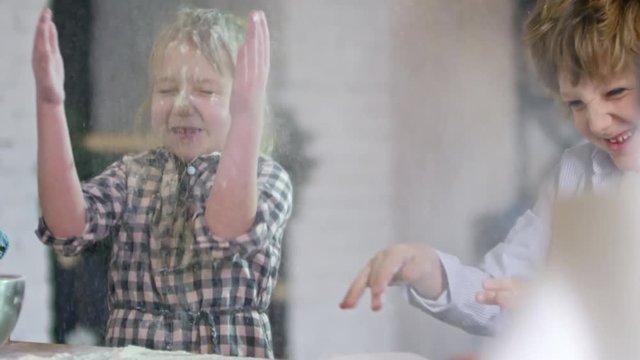 Medium shot of two active kids of elementary school age clapping hands with flour and smiling when cooking in kitchen together