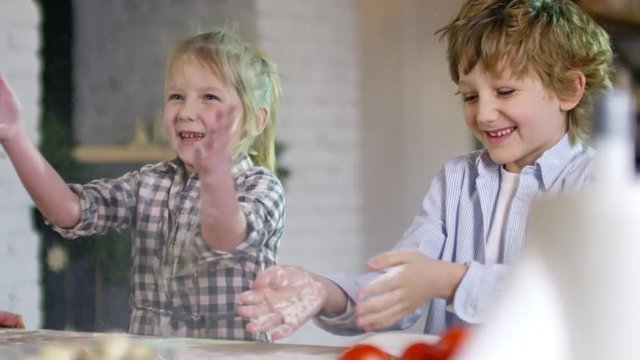 Two adorable little children of primary school age clapping hands full of flour and smiling when having fun in kitchen