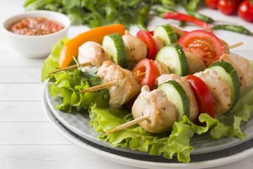 Traditional chicken kebab on wooden skewers with tomatoes, cucumbers and fresh herbs on a plate.