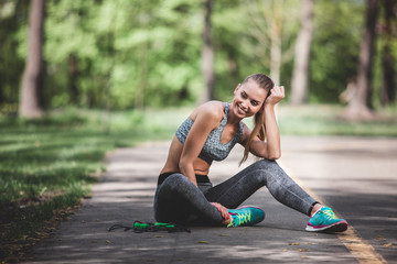 Smiling lady is sitting on road in green countryside. She is having rest during exercising with jumping rope. Pleasant relaxation after work out concept