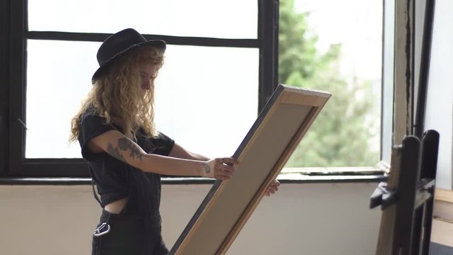 Good-looking painter examining the picture, looking carefully all around the painting near studio window on warm windy morning, wearing black hat and casual outfit