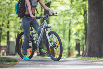Fototapeta premium Low angle of sporty lady riding bike among green landscapes. She is carrying backpack and holding bottle of water. Travelling on bicycle in nature concept