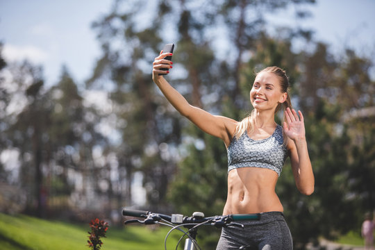 Smiling athletic lady is having fun while cycling in park. She is holding smartphone at arm length both smiling and waving hello to camera. Female is taking snapshot of herself