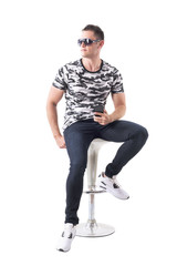 Confident handsome man in camo shirt with sunglasses sitting on bar stool looking away. Full body isolated on white background. 