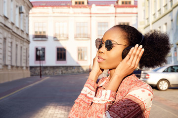 Black afroamerican girl has a walk in evening town and listening to music in headphones.  Girl in sunglasses in summer smiles and looking on sunset.