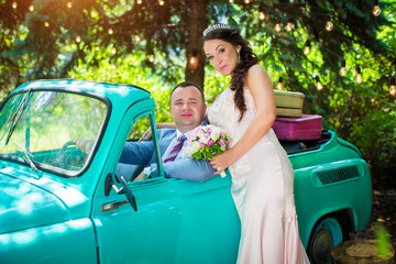 Stylish groom, sitting behind wheel in retro car, and near it stands is charming bride in pink dress, on background of summer park