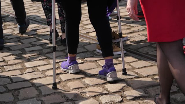Female legs in sneakers walks with help of crutches on a city cobblestone crowd place