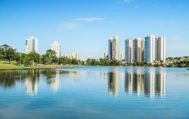Lake of a urban park on a beautiful sunny day. The water of the lake with some buildings on the background and nature around. Photo at Campo Grande MS, Brasil. - obrazy, fototapety, plakaty