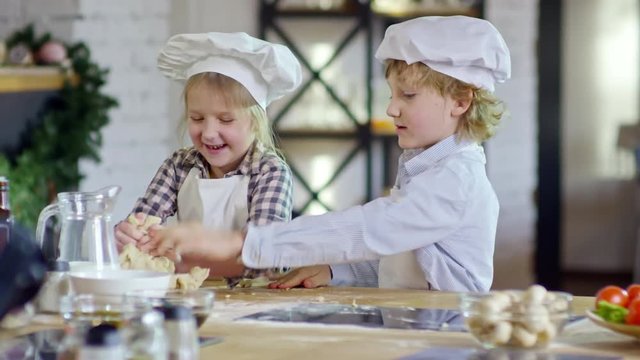 Medium shot of two interested children of elementary school age kneading dough together and enjoying cooking in kitchen