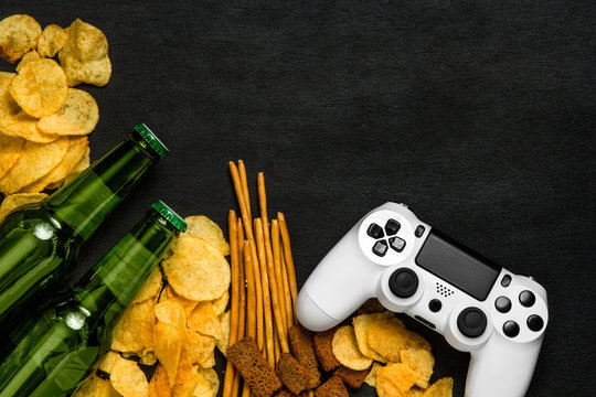Modern white gamepad among chips, crackers and bottles of beer on a black. Game concept.