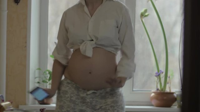 Pregnant woman (with no visible face) in pajama pants and tied white shirt is dancing in front of the kitchen window. Her moves are graceful with some funny elements. She is holding smartphone in the
