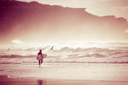 Surfer walking into surf with surfboard under arm at sunrise pink light