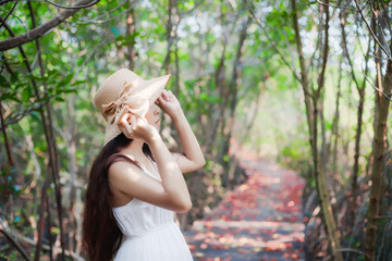 Beautiful asian woman wearing white dress and straw hat enjoy the view with a smile., Standing on a wooden bridge extended into the forest with rays of sunlight beaming through the leaves of the tree.