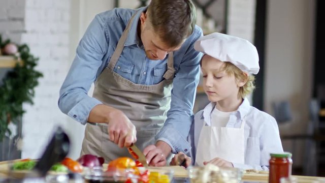 Medium shot of diligent little boy in apron and chef hat watching his father cut pepper on kitchen worktop