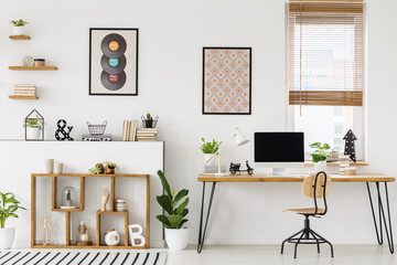 Real photo of a desk with a computer screen standing with a chair next to shelves with ornaments in...