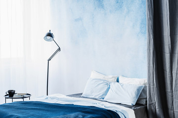 Blue bed between lamp and grey curtain in simple bedroom interior with table and ombre wall