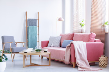 Real photo a pink couch with cushions and blanket standing in a living room next to an armchair and a wooden table with a ladder in the background and windows with blinds