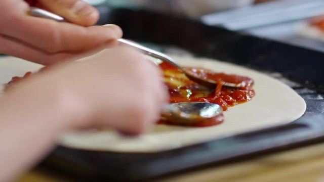 Close up shot of unrecognizable man and child using spoons to apply and spread tomato sauce on pizza dough