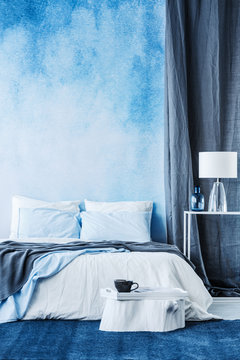 White tree stump with tray and cup of coffee in front of a cozy bed with blue blanket standing against an ombre wall in bedroom interior