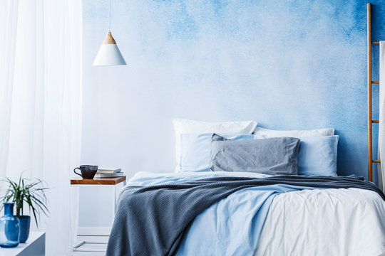 White lamp above table next to bed with blue and grey bedsheets in minimal bedroom interior with plant