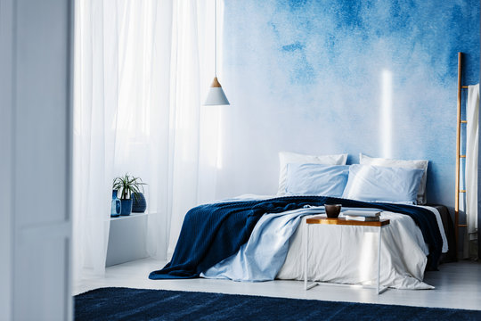 Table in front of bed with navy blue blanket in bedroom interior with ombre wall