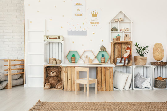 White kid room interior with gold posters on the wall, toys and pillows placed on crate shelves and desk with artificial cactuses