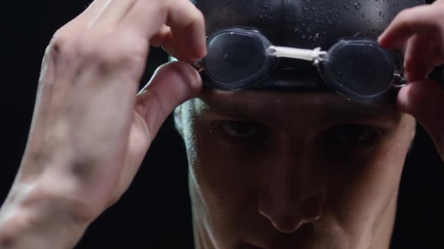 PAN with close up of determined professional athlete in swimming cap stretching isolated on black background and looking at camera, then putting on goggles