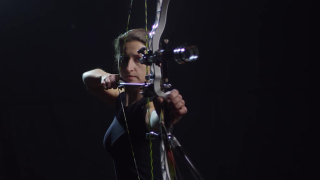 Studio shot of determined female archer with bow shooting arrow against black background