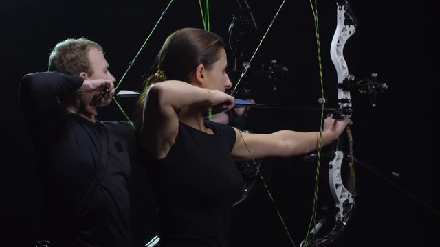 Tilt Up Of Focused Sportswoman And Sportsman With Bows Aiming And Shooting Arrows While Standing Against Black Background