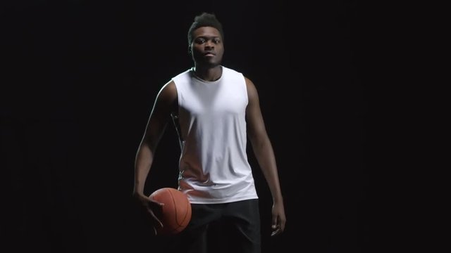 Studio shot with dark background of confident basketball player walking and dribbling ball, then looking at camera and pointing at camera