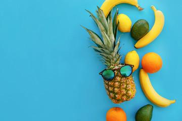 pineapple in stylish sunglasses, and bananas, oranges, avocado and lemons on bright blue paper, trendy flat lay. space for text. fruits modern image, top view. juicy summer background