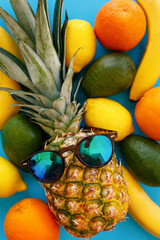 pineapple in stylish sunglasses, and bananas, oranges, avocado and lemons on bright blue paper, trendy flat lay. space for text. colorful fruits modern image, top view. juicy summer pattern