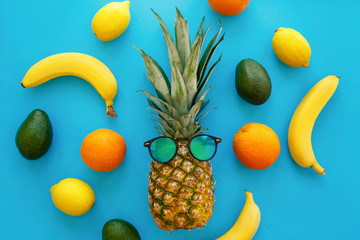pineapple in stylish sunglasses, and bananas, oranges, avocado and lemons on bright blue paper, trendy flat lay. space for text. colorful fruits modern image, top view. juicy summer pattern