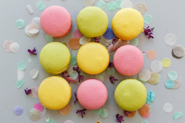 Fototapeta na wymiar colorful macarons on trendy pastel gray paper with lilac flowers and confetti, flat lay. tasty pink, yellow, green and brown macaroons. candy bar for party. food photography