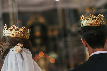 Fototapeta na wymiar bride and groom standing with golden crowns with cross at holy altar during wedding ceremony in church, back view. spiritual moments of holy matrimony