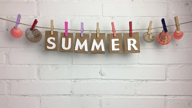 Palabra &ldquo;summer&rdquo; y conchas de mar en una cuerda con pinzas, sobre fondo de ladrillos.