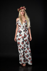 full length portrait of blonde girl wearing floral dress and a flower crown. standing pose against a black studio background.