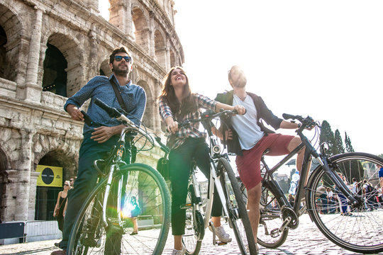 Three happy young friends tourists with bikes and backpacks at Colosseum in Rome taking selfies pictures with smartphone and stick having fun on sunny day