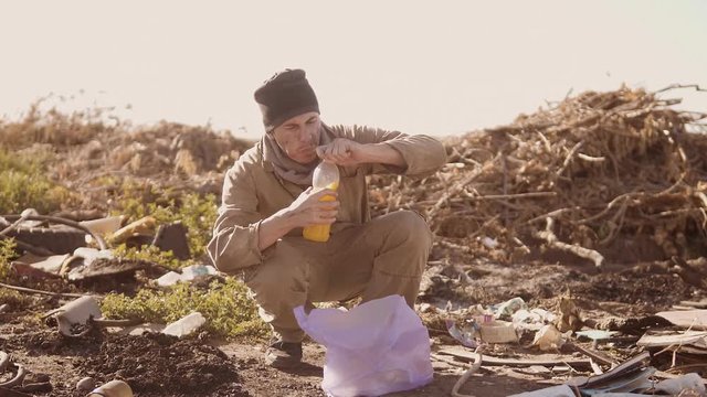portrait of a dirty homeless man in a dump drinks the missing juice looking for food in the package with walking goes looking for food slow motion video. homeless dirty man roofless person looking for