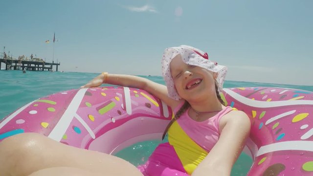 A little girl in a cool hat and a bright bikini is riding on a circle and making a selfie, smiling at the camera